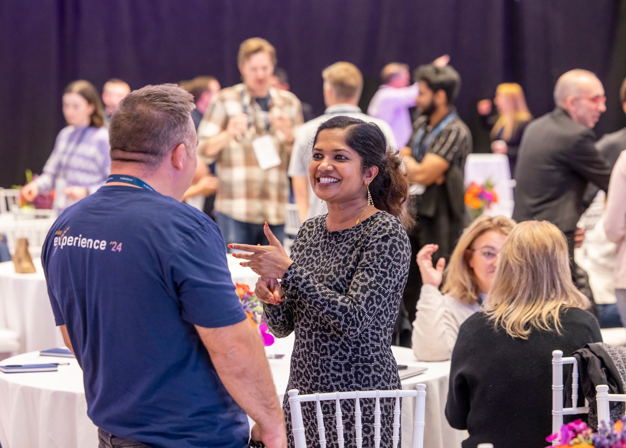 A room of people with two in the foreground who are talking and smiling. The woman is wearing a leopard-print shirt and the man is wearing a navy blue t-shirt. A room of people with two in the foreground who are talking and smiling. The woman is wearing a leopard-print shirt and the man is wearing a navy blue t-shirt.